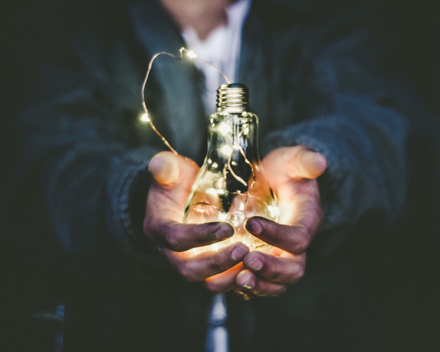 Close-up image of a person holding a lightbulb. The background is dark but the lightbulb is bright and in focus.