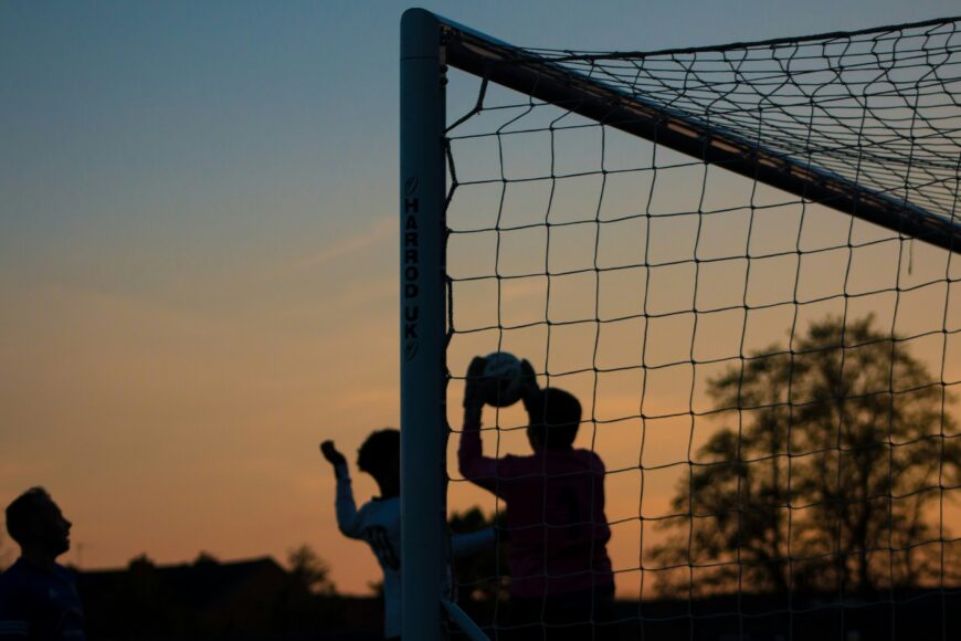A photo of a group of people (blacked out, so only their outlines are visible), playing football in front of a goal. In the background, a sunset with mustard yellow and sky blue tones.